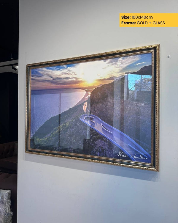 Framed artwork of a scenic landscape with a sunset over water, displayed on a wall.