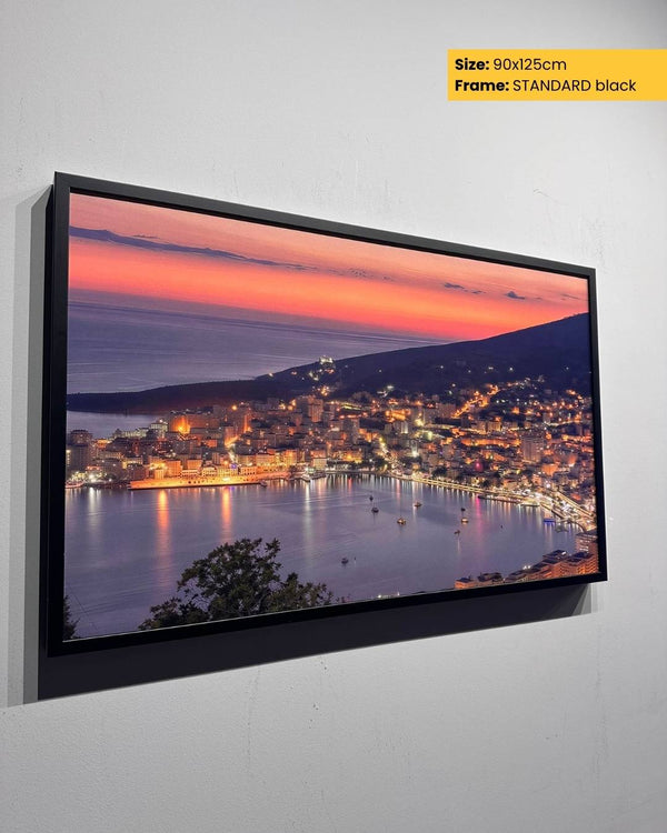 Framed artwork of a cityscape at dusk with a body of water, on a white wall.