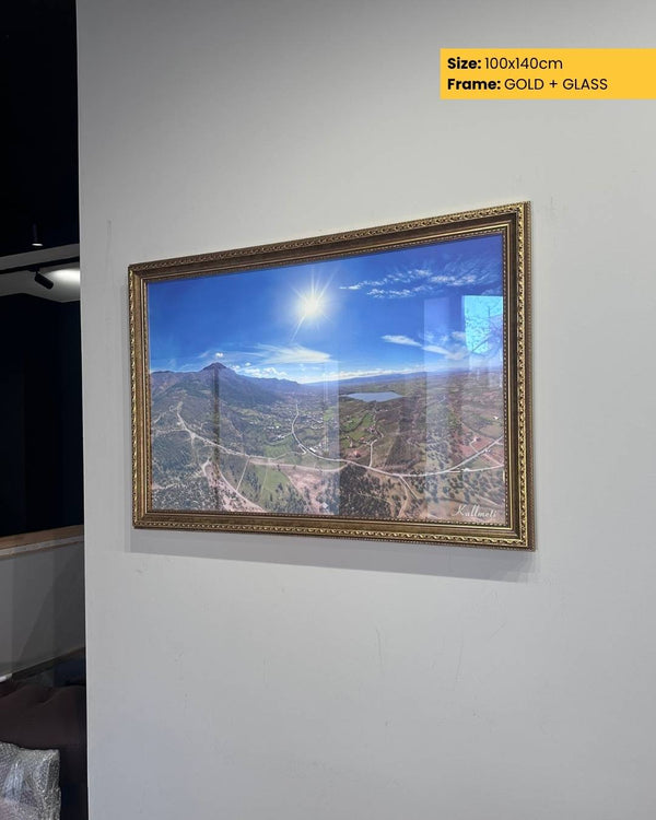 Framed artwork of a mountain landscape with gold frame and glass on a white wall.
