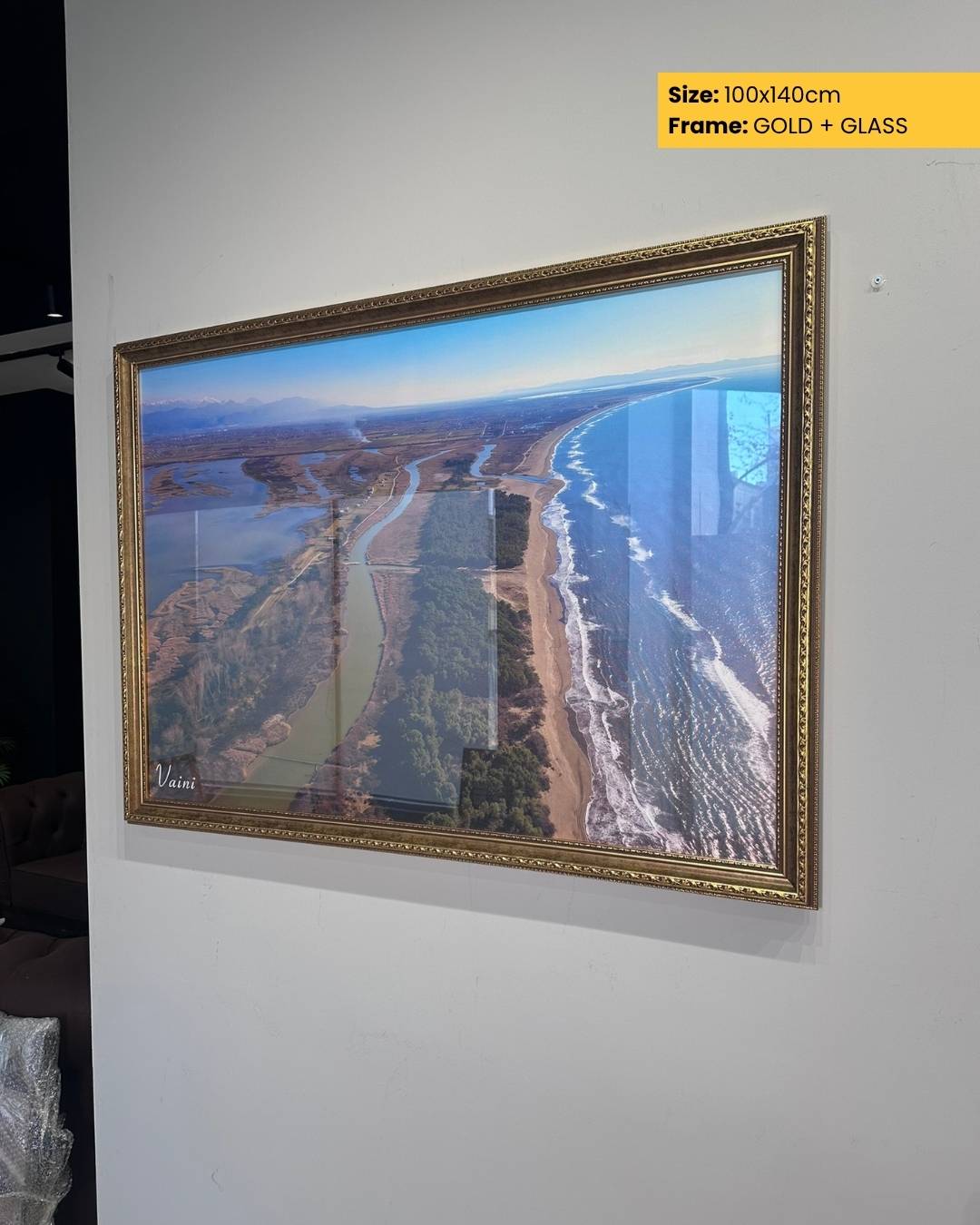 Framed artwork of a landscape with a river and buildings, displayed on a wall.
