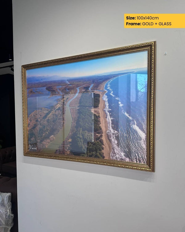 Framed artwork of a landscape with a river and buildings, displayed on a wall.