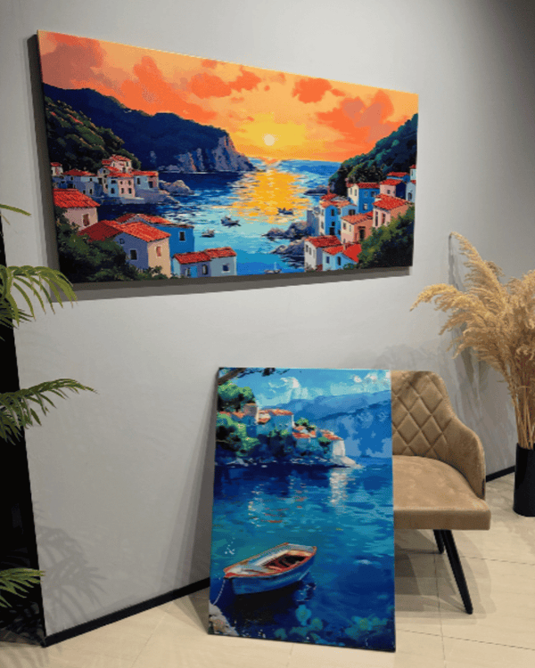 Two artworks of a coastal scene with houses and boats, one on a wall and one on a floor.