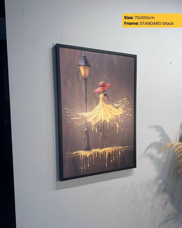 Framed artwork of a woman in a yellow dress and red hat under a street lamp on a white wall.