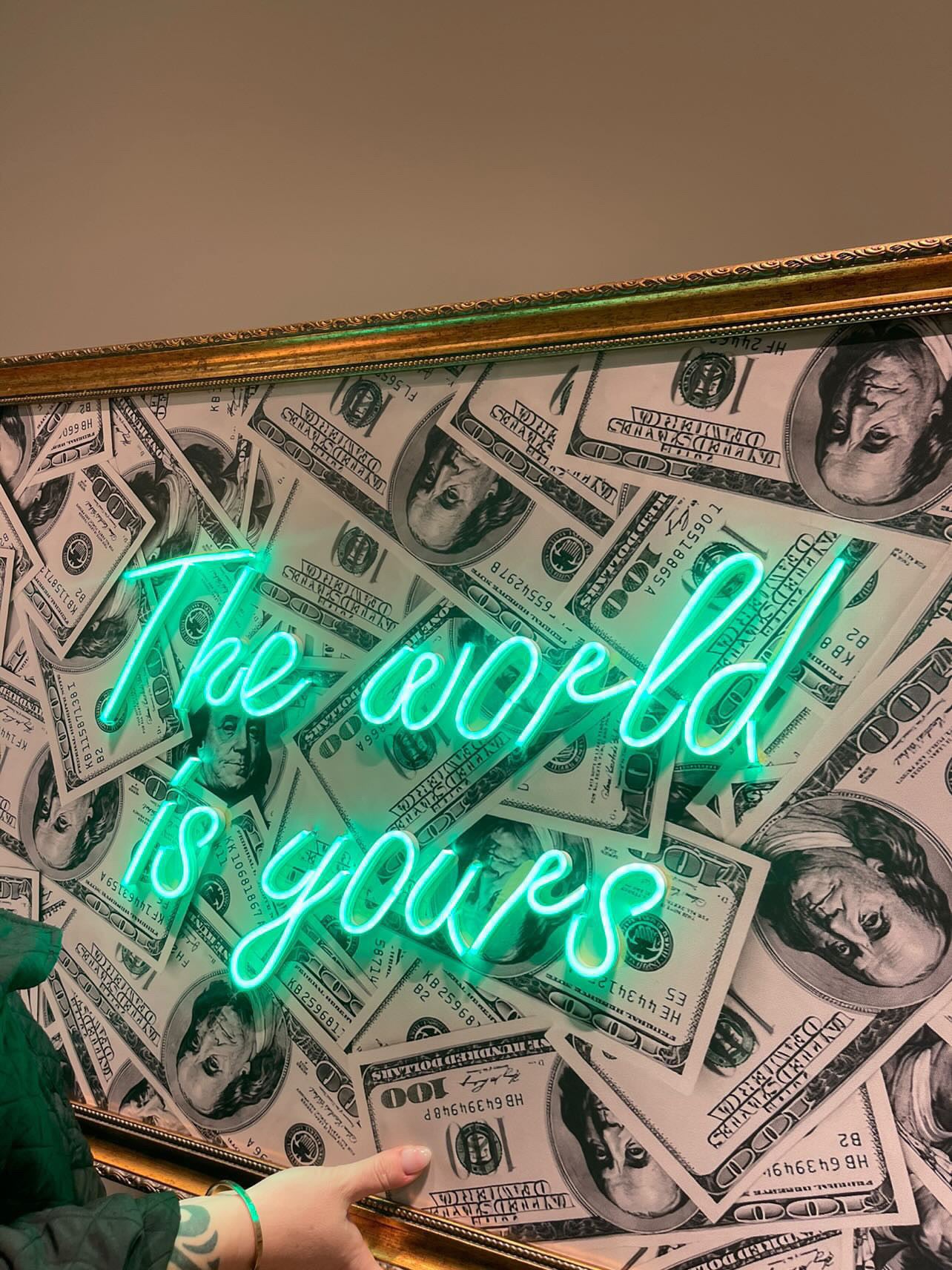Neon sign with 'The world is yours' on a background of dollar bills