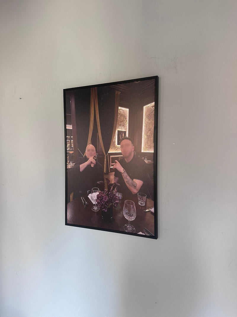 Framed photograph of a person sitting at a table with drinks on a white wall.