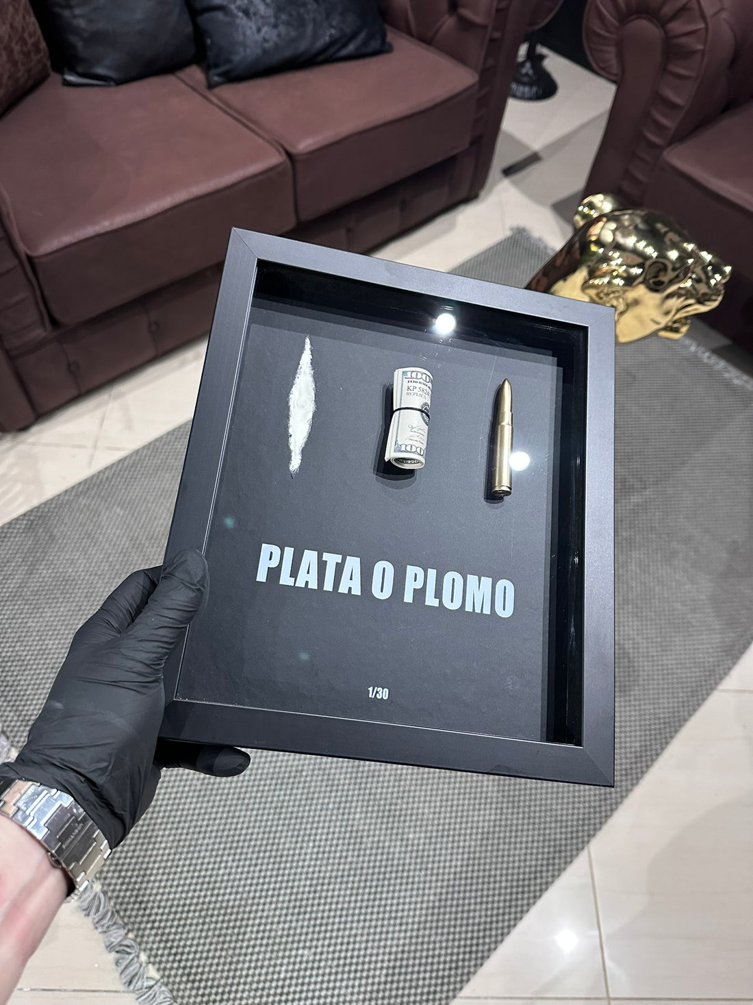 Black shadow box with 'PLATA O PLOMO' text held by a person in a room.