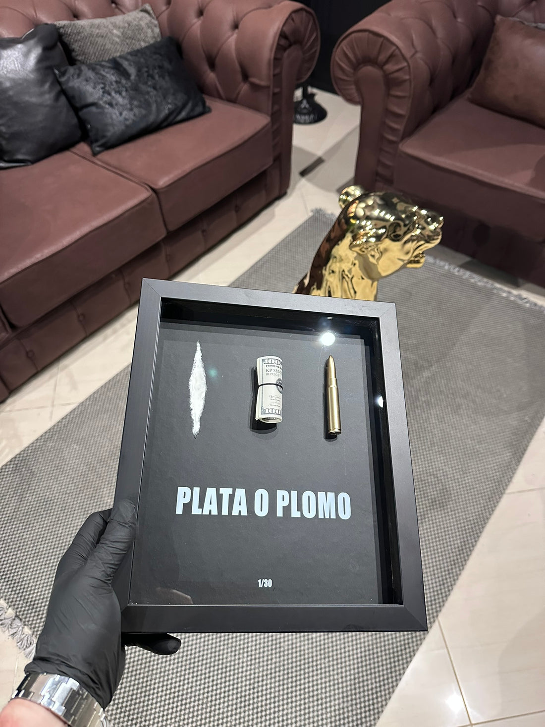Black shadow box with 'PLATA O PLOMO' text held by a person in a room.