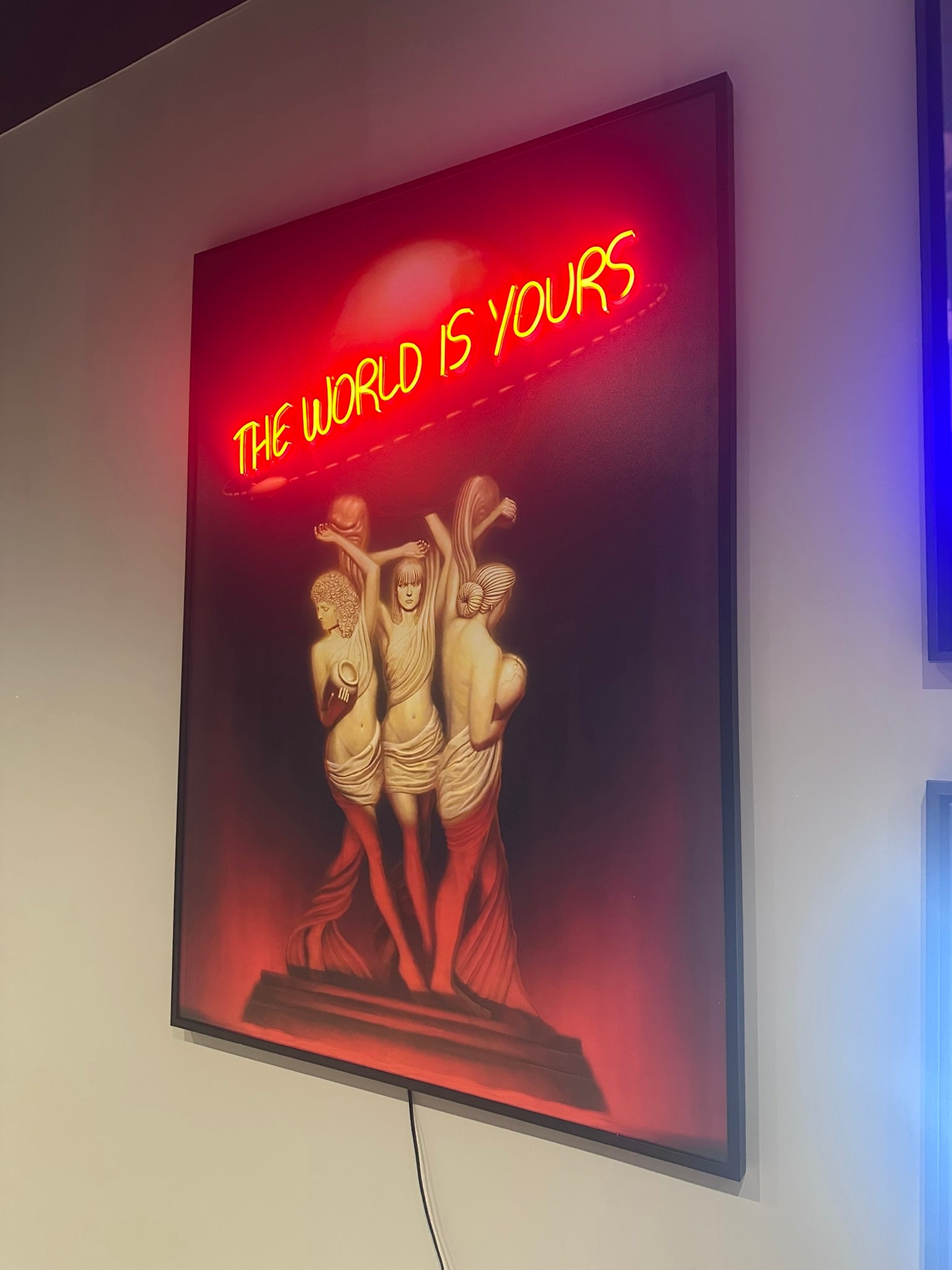 Neon sign with 'The World is Yours' text above classical sculpture replica on a red background.