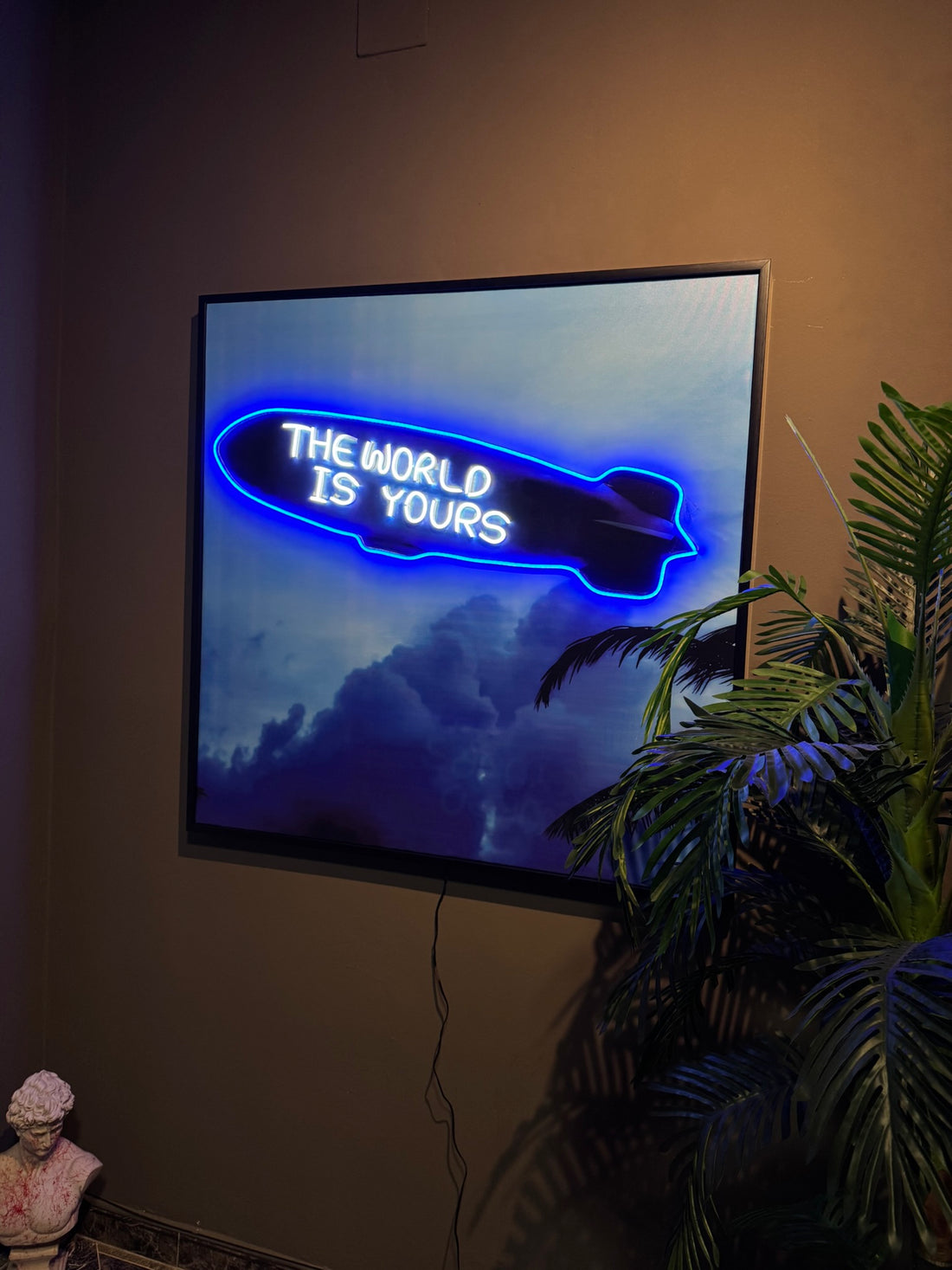 Neon sign on a wall with 'The World is Yours' text in a dark room with a plant.
