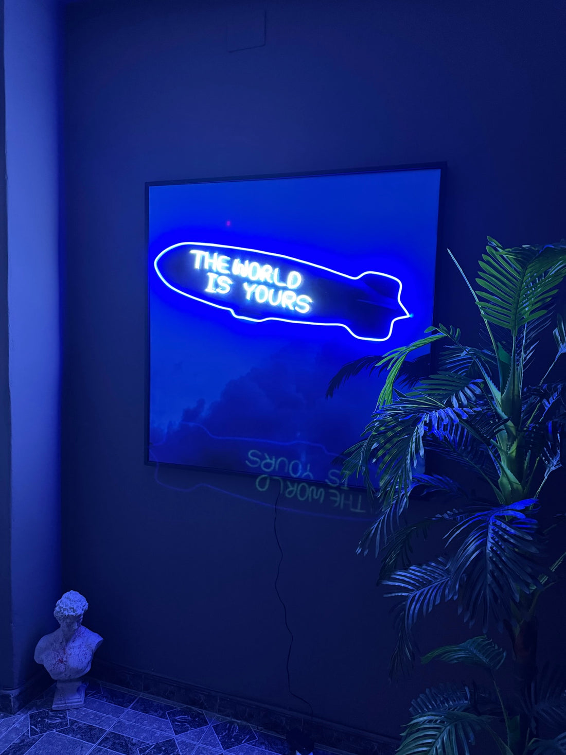 Neon sign on a wall with 'The World is Yours' text in a dark room with a plant.