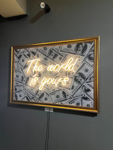 Neon sign with 'The world is yours' text on a dollar bill patterned background, framed in gold against a dark wall.