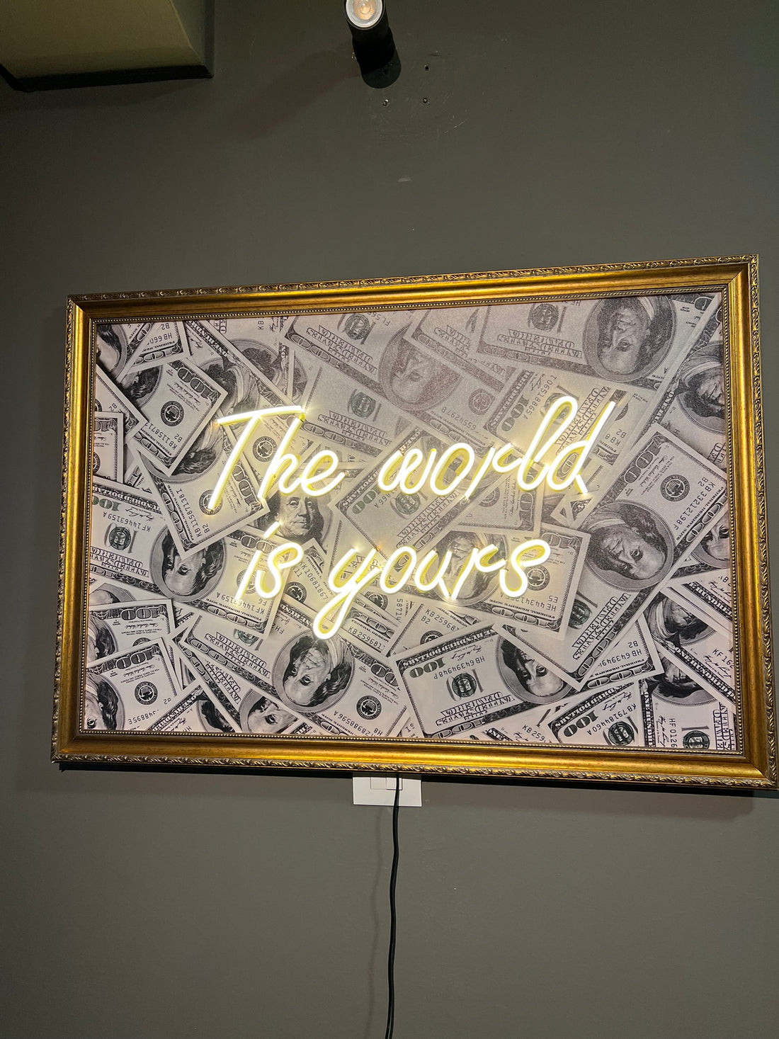 Neon sign with 'The world is yours' text on a dollar bill patterned background, framed in gold against a dark wall.