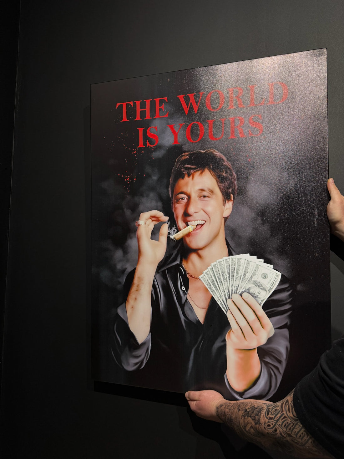 Person holding a poster with a man smoking and money, text 'The World is Yours' on a dark background