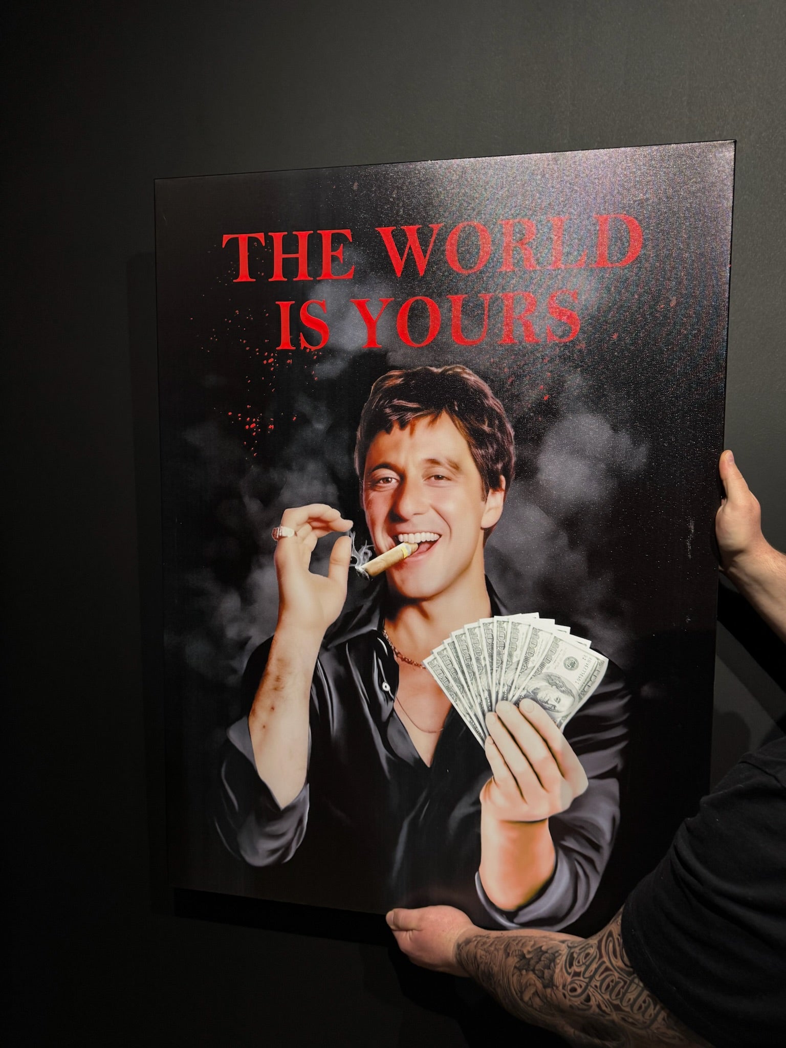 Person holding a poster with a man smoking and money, text 'The World is Yours' on a dark background