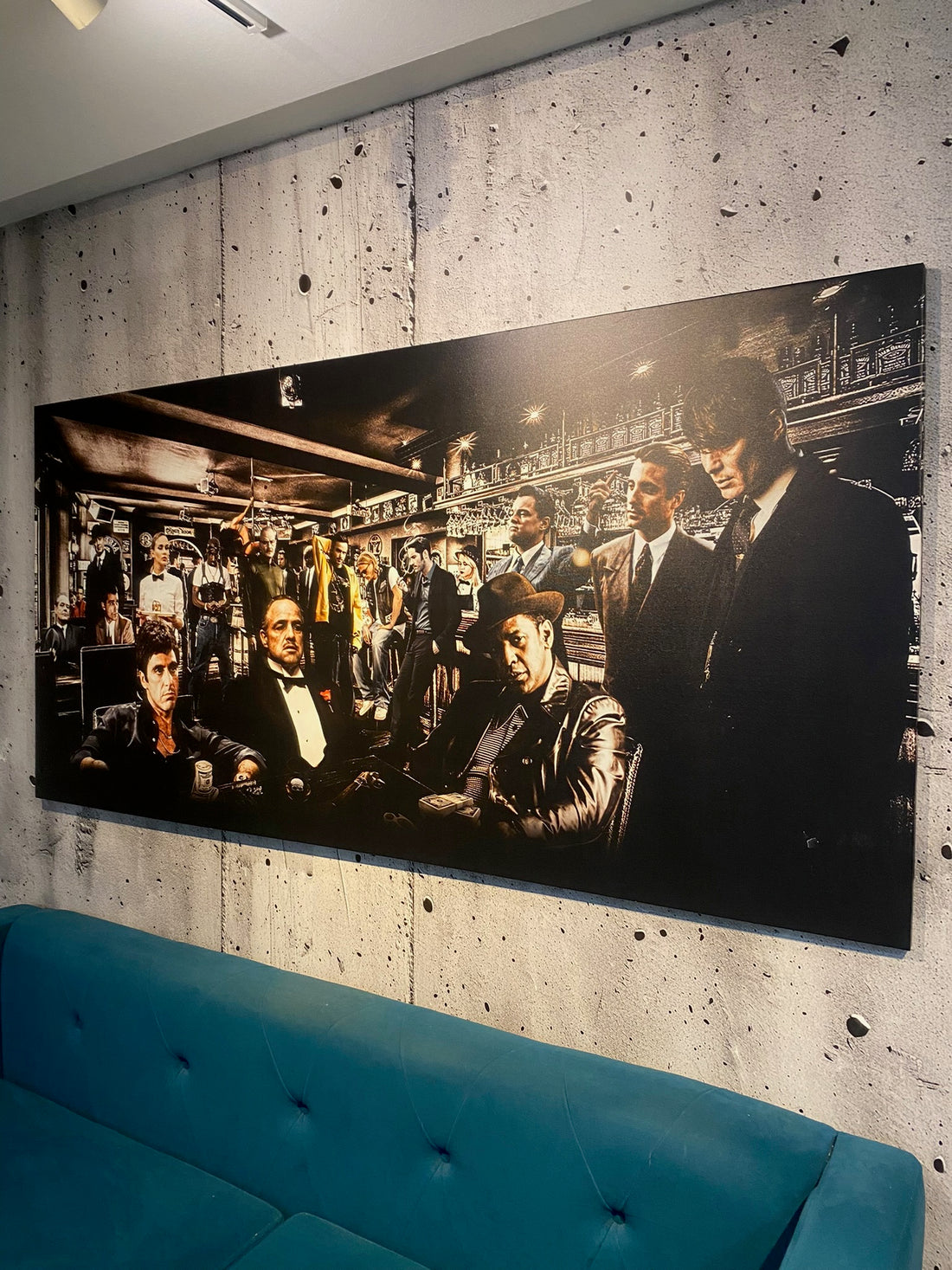 Large black and white painting of a crowded room on a concrete wall above a blue sofa.