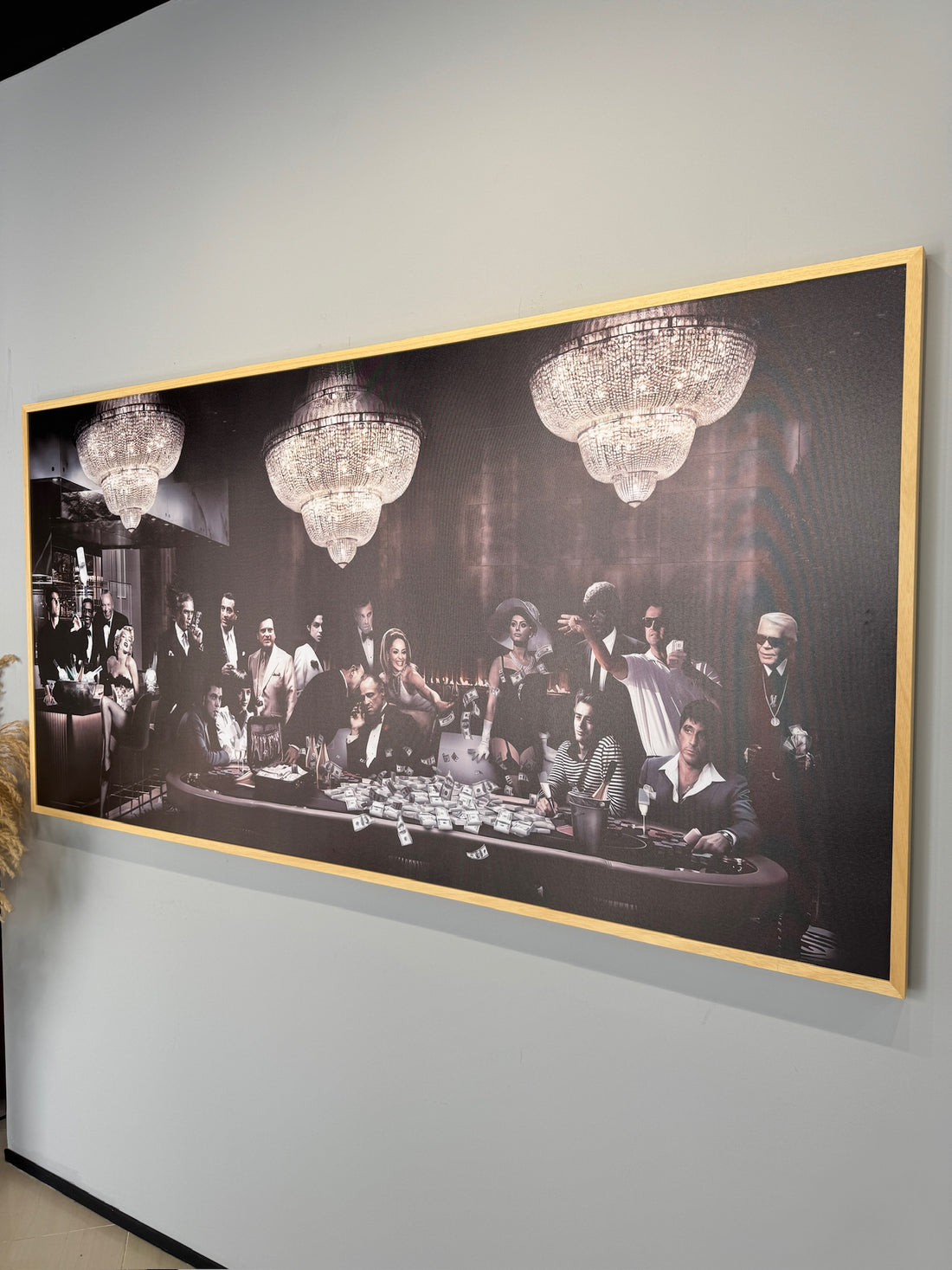 Framed black and white artwork of a group of people around a table with chandeliers above, on a gray wall.