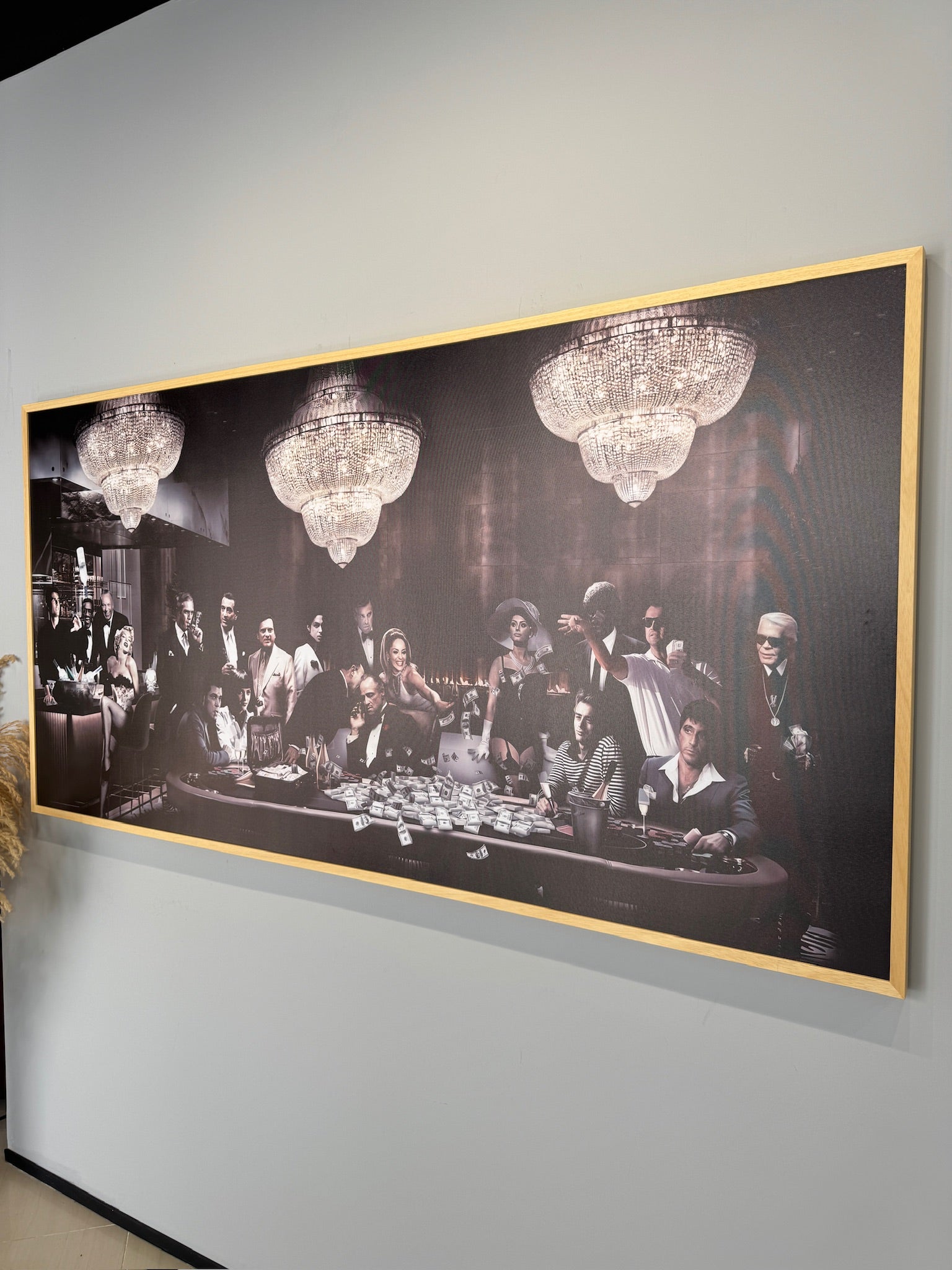 Framed black and white artwork of a group of people around a table with chandeliers above, on a gray wall.