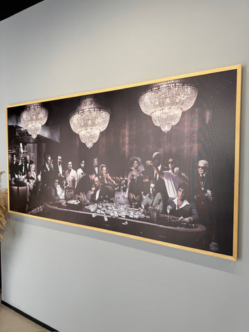 Framed black and white artwork of a group of people around a table with chandeliers above, on a gray wall.
