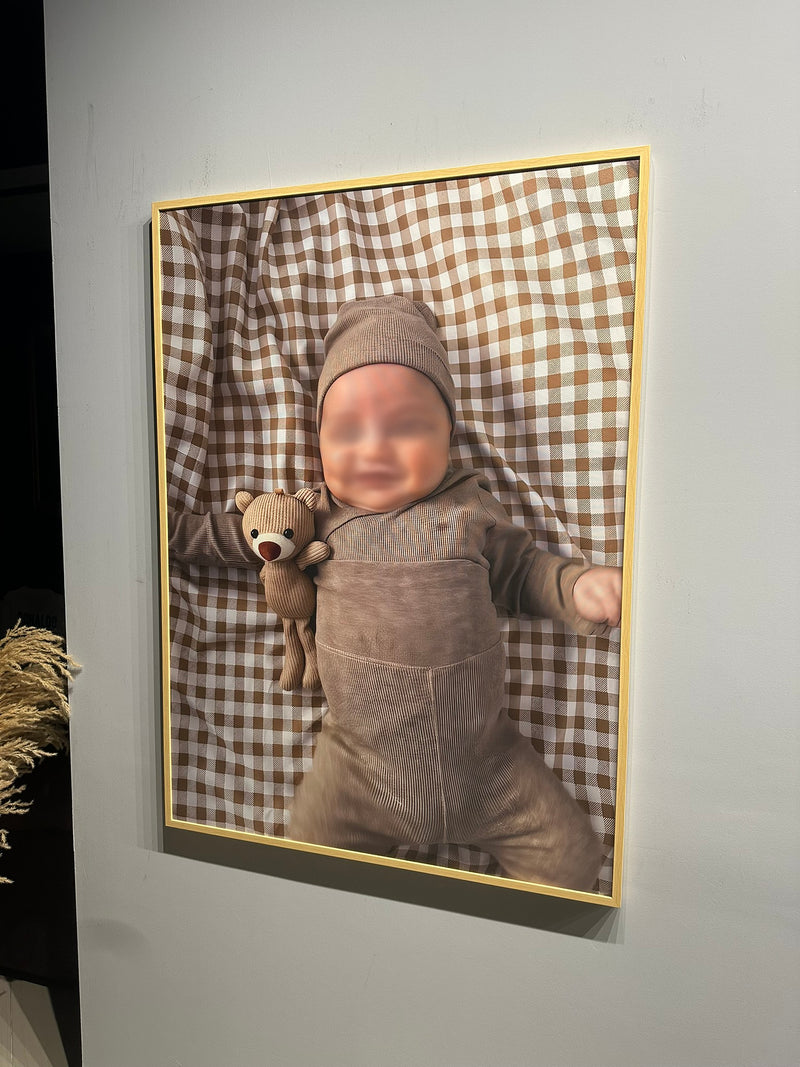 Framed picture of a baby in a brown outfit holding a teddy bear against a checkered background.