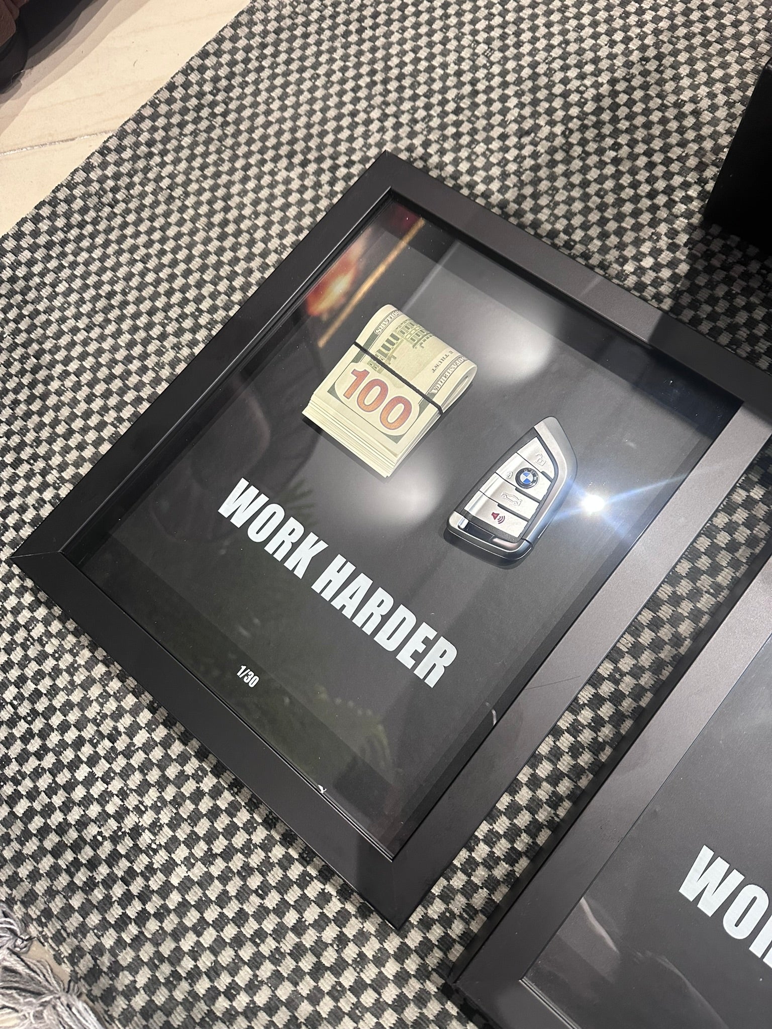 Framed motivational quote 'Work Harder' with a car key and 100 dollar bill on a checkered fabric background.